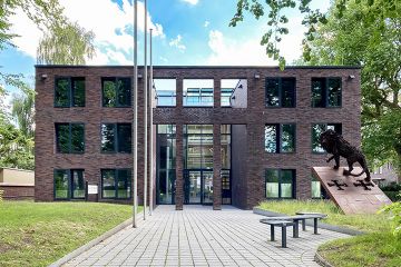 Ein kastiges Gebäude aus modernem lila-braunem Ziegelstein, schwarzen Fenstern und einem gläsernen Eingang